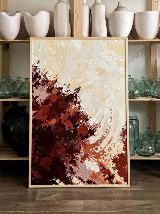 A modern artwork piece with a blend of red and maroon hues on a light wooden frame, displayed in an home setting with various decorative items in the background.